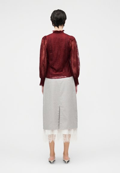 Bordová čipková blúzka s nafúknutými rukávmi, skombinovaná so sivou midi sukňou s rozparkom na chrbte. Biela čipkovaná lem viditeľná pod sukňou.