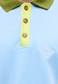 Polo bleu clair avec col vert olive, trois boutons marron et motif floral brodé blanc sur la poitrine.