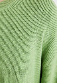 Grønn strikket genser med teksturert finish, ribbede detaljer og bred hals. Stoffet virker mykt og lett.