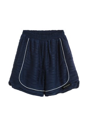 Joven con pantalones cortos texturizados azul marino y camiseta con etiqueta, calcetines blancos y zapatos beige sin cordones, de pie frente a un fondo blanco.