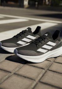 Baskets de sport noires avec une tige en maille texturée, dotées de trois rayures blanches et d'une semelle blanche épaisse. Les accents incluent un logo sur le talon.