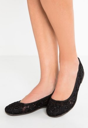 Stopy noszące czarne koronkowe baletki z kwiatowym wzorem, stojące na białej powierzchni.