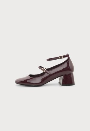 Chaussure en cuir verni violet foncé à talon moyen, avec deux brides réglables à la cheville et un bout arrondi sur un fond clair uni.