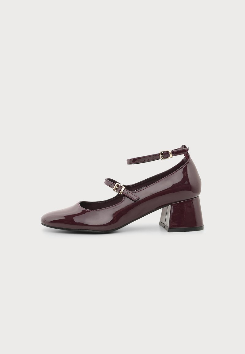 Chaussure en cuir verni violet foncé à talon moyen, avec deux brides réglables à la cheville et un bout arrondi sur un fond clair uni.