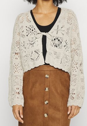 Cardigan en tricot beige clair avec un motif ajouré, design court, manches longues et fermeture par lien à l'avant. Porté sur un haut noir et une jupe marron.
