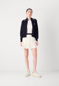 Tamsiai mėlynas zip-up švarkas su raudonais akcentais, dėvimas su baltais prigludusiais marškiniais ir balta plisuota sijonu. Avalynė - šviesių tonų sportbačiai.