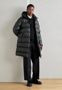 Lange zwarte pufferjas met een capuchon, voorzien van horizontaal gestikte delen, voorzakken met rits en een ontspannen pasvorm. Draag het over een donkere outfit.