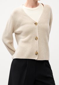 Hellbeige gestrickte Strickjacke mit V-Ausschnitt, fünf großen sandfarbenen Knöpfen und leicht verkürztem Schnitt. Strukturierter Stoff mit gerippten Details.
