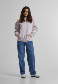 Haut à capuche rose clair avec une poche frontale et un cordon de serrage, associé à un jean en denim bleu ample et des baskets blanches avec des rayures noires.