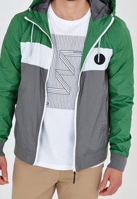 Zielona, szara i biała lekka kurtka z kolorowymi blokami, z kapturem, zapinana na zamek, z elastycznymi mankietami i okrągłą naszywką z logo. Pod nią znajduje się biała koszulka z grafiką.