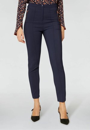 Kvinde iført marineblå, slanke ankellange bukser med frontsøm, lynlåslommer i siden og sorte spidse hæle, kombineret med en blomstret langærmet top.