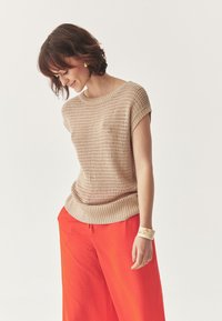 Maglione a maniche corte lavorato a maglia beige con orlo a coste e trama intricata, abbinato a pantaloni larghi rossi vivaci e un braccialetto di colore chiaro.