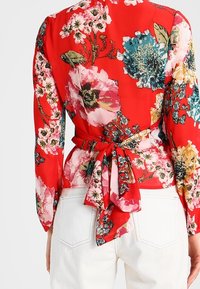 Blusa rossa floreale con grandi fiori rosa, blu e verdi, maniche lunghe e dettaglio con fiocco in vita sul retro.