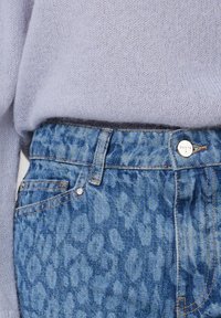 Vervaagde blauwe jeans met een luipaardprintpatroon, met een hoge taille, een zilveren knoop en een zacht lavendelkleurig truitje erboven.