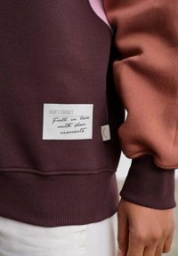 Violetinė ir rožinė džemperio su tekstūruota etikete šone, kurioje parašyta "Neužmiršk" ir "Įsimylėk lėtas akimirkas."