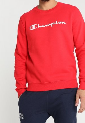 Mand iført en rød Champion sweatshirt og marineblå Russell Athletic joggingbukser, stående med den ene hånd i lommen.