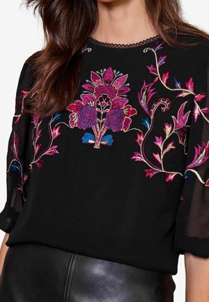 Černá průsvitná halenka s růžovou, modrou a zlatou květinovou výšivkou na hrudi a rukávech, nošená s černou koženou sukní.
