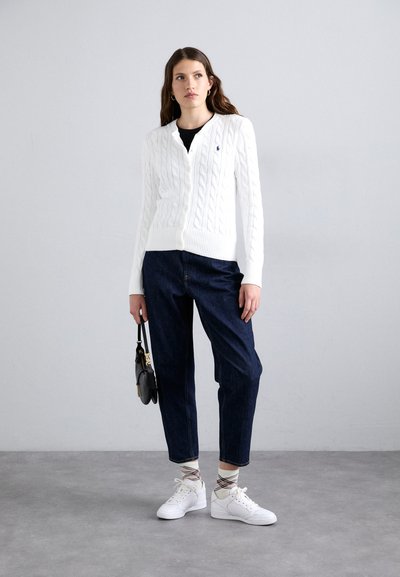 Weißer Strickcardigan mit Knöpfen, kombiniert mit dunklen Jeanshosen. Weiße Sneakers und gemusterte Socken. Das Model hält eine kleine schwarze Handtasche.