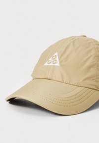 Cappello beige con una toppa a forma di triangolo bianco. Realizzato in tessuto leggero con dettagli cuciti lungo il visiera e la corona.