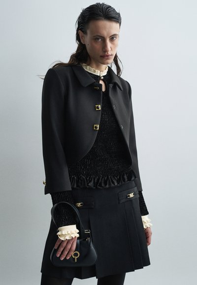 Black cropped jacket with gold button accents, textured black top with ruffled hem, and a black purse with a keyhole detail.