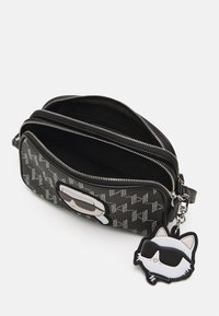 Mustan keinonahkainen laukku, jossa on geometrinen kuosi, hopeiset vetoketjut, kaksi osiota ja irrotettava kissanmuotoinen avaimenperä, jossa on aurinkolasit.