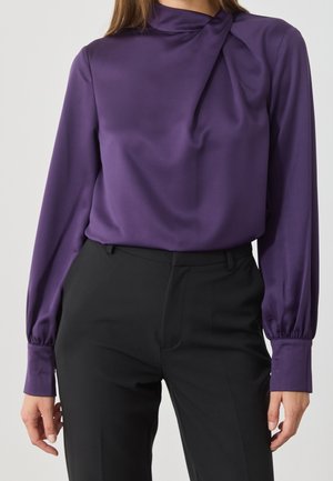 Kvinde iført en lilla langærmet satinbluse med draperet halsudskæring, stukket ned i højtaljede sorte skræddersyede bukser.