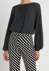 Blusa grigia a fascia ampia con dettagli plissettati, abbinata a pantaloni a vita alta a righe bianche e nere con linee diagonali.