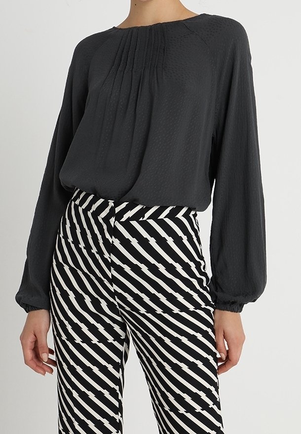 Blusa grigia a fascia ampia con dettagli plissettati, abbinata a pantaloni a vita alta a righe bianche e nere con linee diagonali.