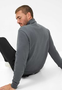 Felpa grigia con colletto alto e schiena textured. Modello seduto, indossando pantaloni neri e scarpe bianche. Tessuto morbido con vestibilità comoda.