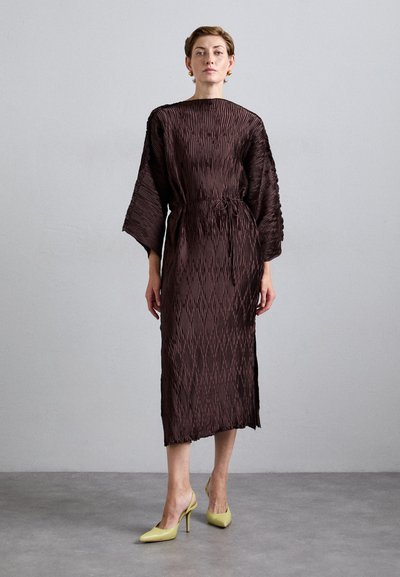 Woman stands against plain gray wall wearing a textured dark brown midi dress with wide sleeves and yellow high-heeled slingback shoes.
