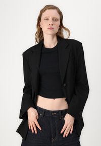 Črn oversized blazer z izrezanimi lapelami, dvema gumboma in žepom. Občan je preko črnega kratkega topa in temno modrih kavbojkah z visokim pasom.