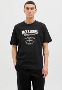 Juodas medvilninis marškinėliai su apvalia iškirpte, trumpomis rankovėmis, drąsiu baltu tekstu ir retro logotipu, atsipalaidavusio kirpimo.
