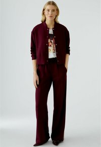 Burgunderfarbene cropped Jacke mit Druckknöpfen, kombiniert mit einem weißen Grafik-T-Shirt und passenden weiten Hosen. Glatte Textur, lässige Passform.