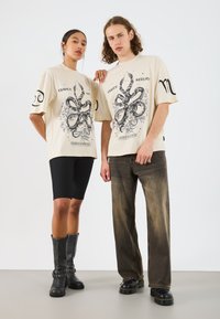 Beige oversized trička s černým grafickým designem, spárovaná s černými cyklistickými šortkami a černými kozačkami; tmavé džíny a robustní boty na modelu.