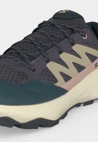 Sötétszürke és zöld sportcipő bézs "N" logóval, vastag fehér talppal és mintás fűzőkkel, elölnézetből, oldalról ábrázolva.