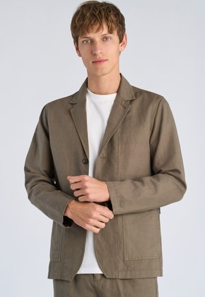 Jonge man die een knoop aanpast op een olijfgroene casual blazer over een wit T-shirt tegen een effen lichte achtergrond.