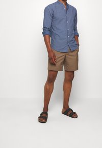 Mies joka on pukeutunut siniseen kuviolliseen pitkähihaiseen paitaan, vaaleanruskeisiin shortseihin ja mustiin solkisaappaisiin, seisoo yksinkertaista valkoista taustaa vasten.