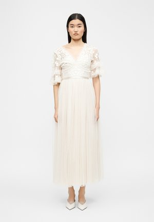 Nő áll egyenesen, hosszú fehér ruhát visel, amelynek virágos csipkebetétes a felső része, áttetsző puffadt ujjakkal, fehér hegyes orrú magassarkú cipővel párosítva.