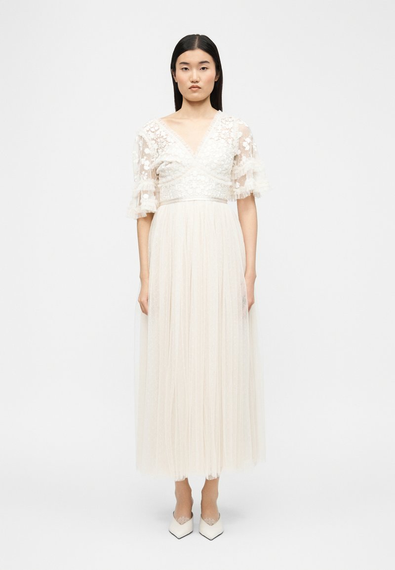 Γυναίκα που στέκεται όρθια φορώντας ένα μακρύ λευκό φόρεμα με κορσέ από δαντέλα με λουλούδια και διαφανή φουσκωτά μανίκια, συνδυασμένο με λευκά μυτερά τακούνια.