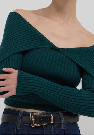 Personne portant un pull côtelé vert foncé à épaules dénudées avec des manches longues et un jean bleu foncé avec une ceinture noire et une boucle dorée.