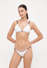 Hvit bikinisett med en trekantet bralette med svarte detaljer og tynne justerbare stropper; matchende lavtsittende bunn med minimal dekning.