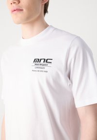 Hvit bomulls t-skjorte med svart logo og tekst på venstre brystområde. Crew neck design med korte ermer og glatt tekstur.