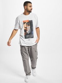 Hvid t-shirt med et grafisk print, der viser et ansigt og tekst, parret med lysegrå cargo bukser og hvide sneakers. Casual pasform og stil.