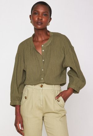 Chemise rayée vert olive avec un col rond et des boutons à l'avant, associée à un pantalon beige taille haute avec des boutons décoratifs.