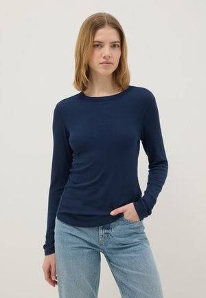Vrouw met schouderlang haar, gekleed in een nauwsluitend marineblauw shirt met lange mouwen en lichtblauwe jeans, staand met één hand in haar broekzak.