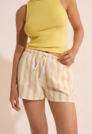 Personne portant un débardeur côtelé jaune et un short blanc avec des rayures verticales jaunes et roses, ajustant le short à taille élastique et cordon.