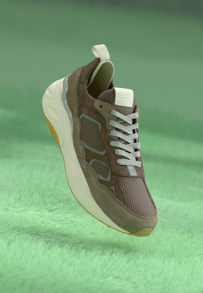 Brown suede sneaker with mesh detailing, gray accents, and white laces. Features a padded tongue and a textured rubber sole.