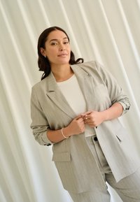 Světle šedý pruhovaný blazer a odpovídající kalhoty přes bílé tričko. Mezi charakteristiky patří klopové límce, kapsy a knoflíkové detaily.