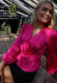 Bluza iz fuksijastega satena z dolgimi napihnjenimi rokavi, ovijajočim dizajnom in vozlom v pasu. Kombinirana s črno oprijeto krilo. Poudarjena z detajlnimi nakitnimi dodatki.