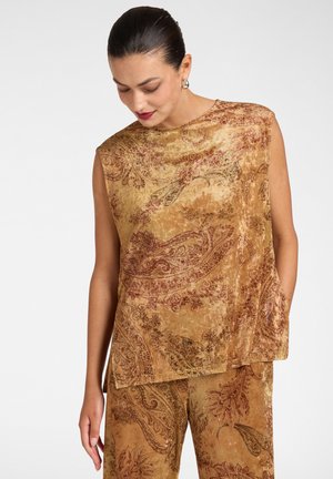 Frau trägt ärmelloses braunes Samtoberteil und passende Hose mit komplizierten Paisley- und Blumenmustern, schaut nach unten, Haare zurückgebunden.